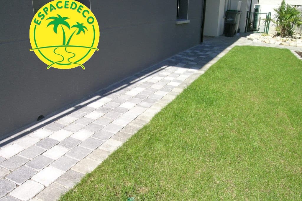 Allée pavée gris et blanc le long d'un mur, bordant une pelouse verte. Logo ESPACEDECO avec palmiers.